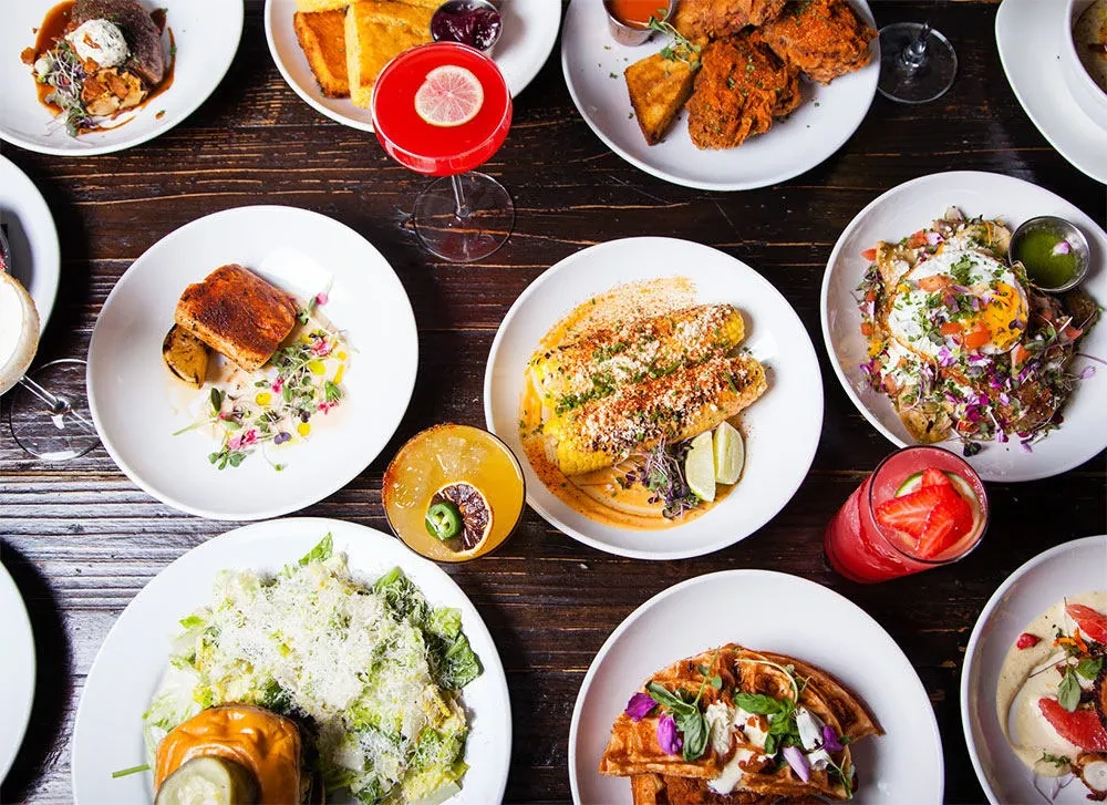 Bright overhead shot: diverse food dishes, colorful cocktails on dark table.