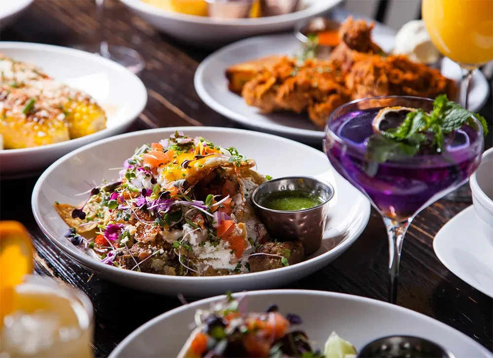 Vibrant brunch spread with chilaquiles, fried egg, purple cocktail, and elote.