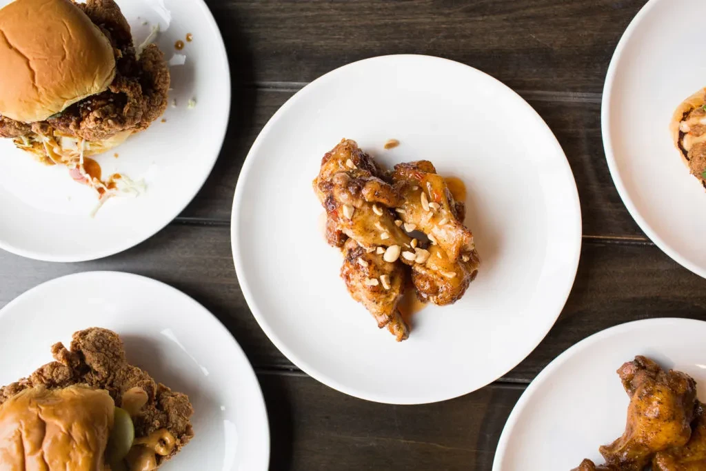 Overhead: Glazed chicken wings and fried sandwiches on a dark table.