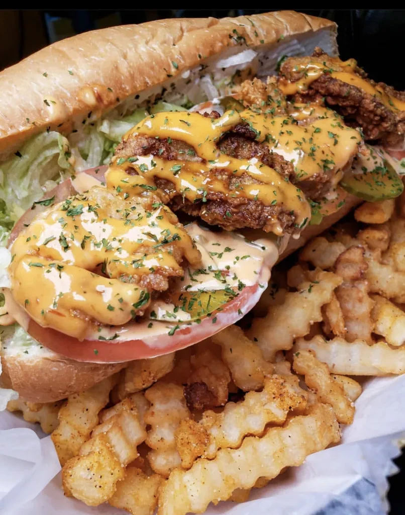 Overflowing fried seafood sandwich with sauce, herbs, and crinkle-cut fries.
