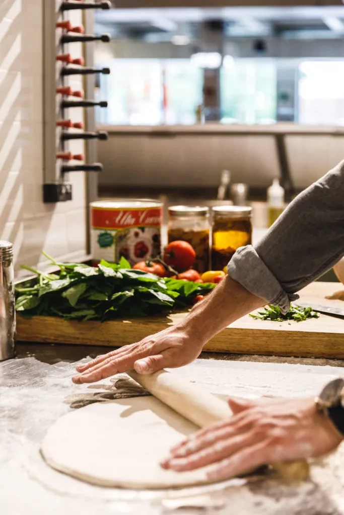 Hands rolling pizza dough on floured surface, fresh ingredients nearby.