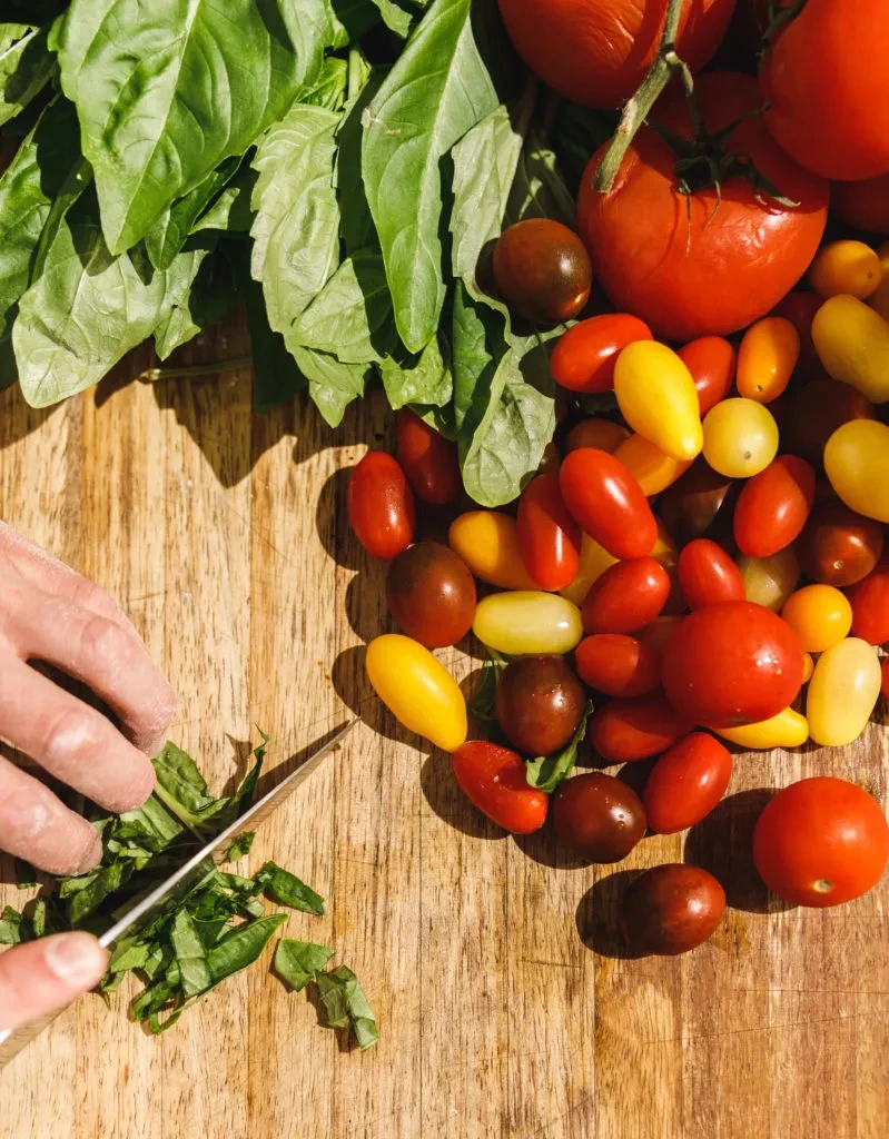 Hand chopping fresh basil and colorful tomatoes on a wooden board.