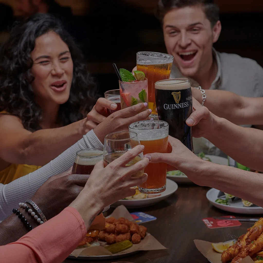 Seven hands clink various drinks in a lively restaurant toast.