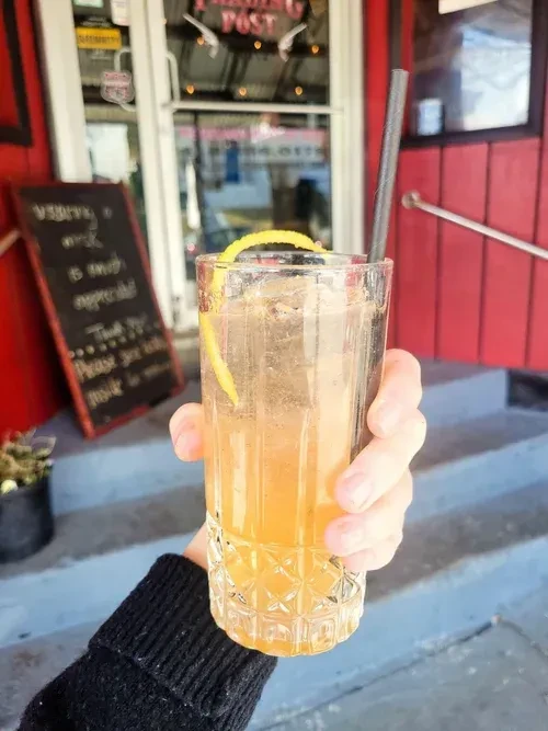 Hand holding an amber drink with ice, lemon peel, and straw outdoors.