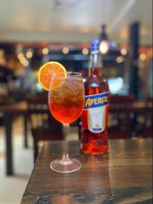 Aperol Spritz cocktail with ice and orange, next to an Aperol bottle.