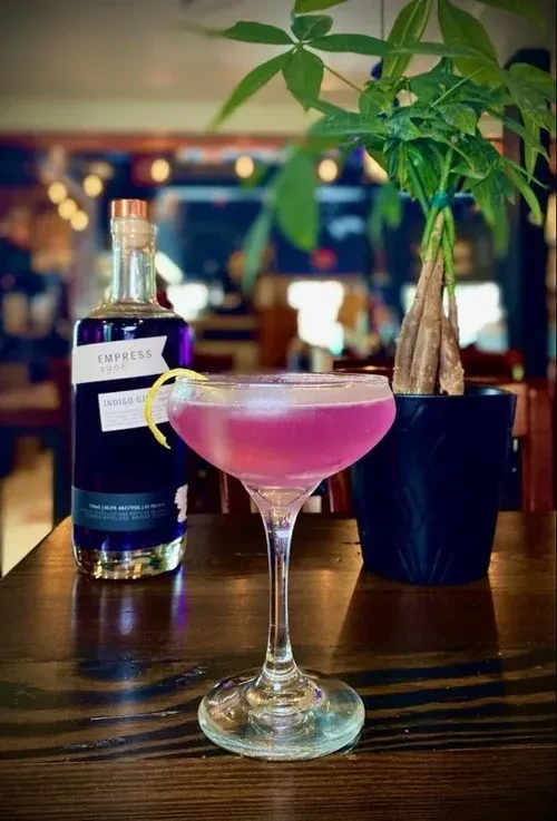 Pinkish-purple cocktail, Empress Gin bottle, and potted plant on a table.