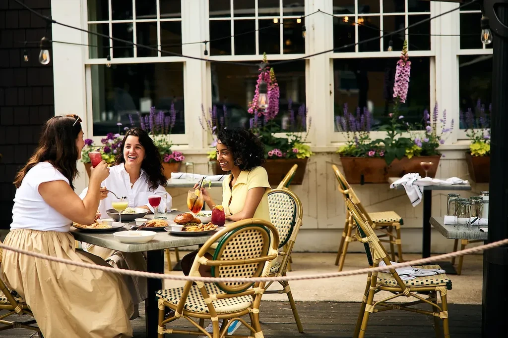 Three women dine outdoors, smiling amidst flowers and string lights.