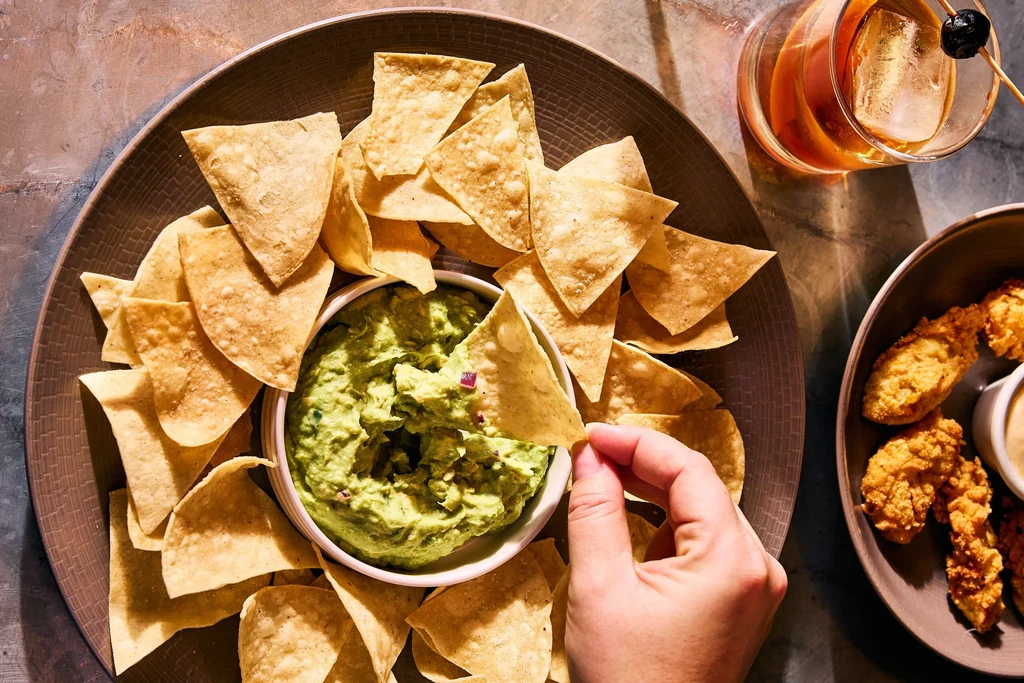 Top-down: Hand dips chip in guacamole; platter, cocktail, and fried appetizers.