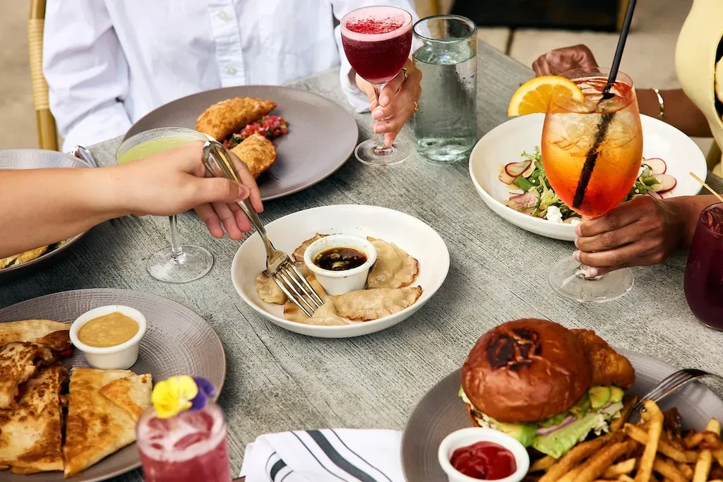 Overhead view of a dining table with diverse food, cocktails, and hands.