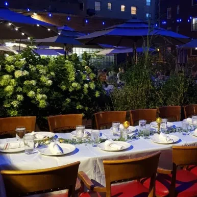 Outdoor dining table set at dusk with foliage and string lights.