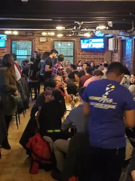 Lively, crowded restaurant interior with visible patrons, TVs, and brick walls.