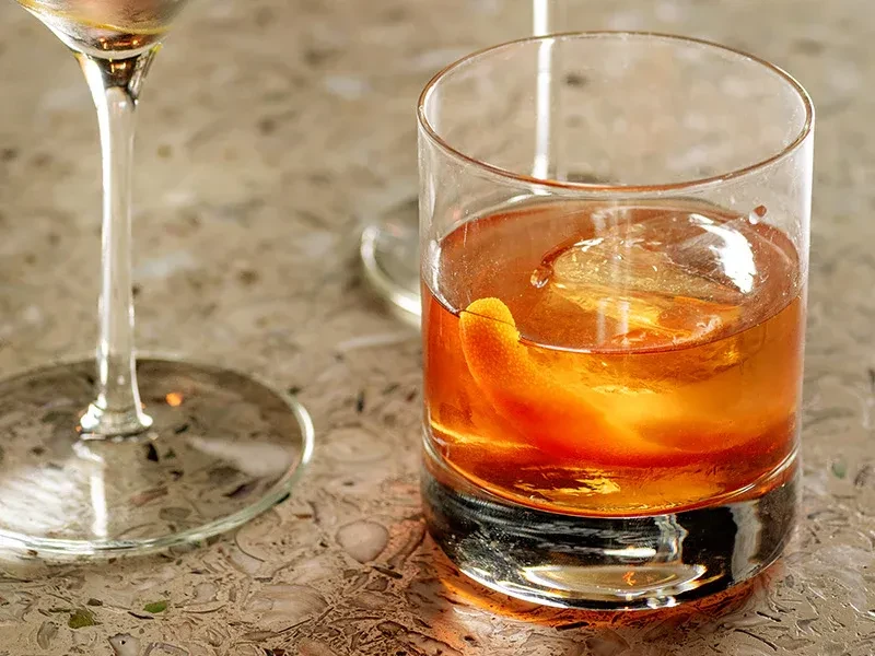 Amber cocktail in rocks glass with large ice cube and orange twist.