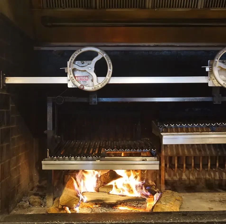 Open-fire grilling station with burning logs in a brick fireplace.