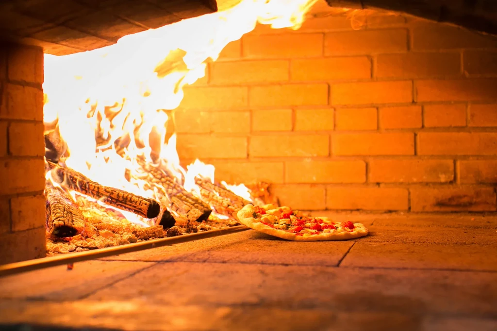Partially cooked cheese pizza in a brick wood-fired oven, next to flames.