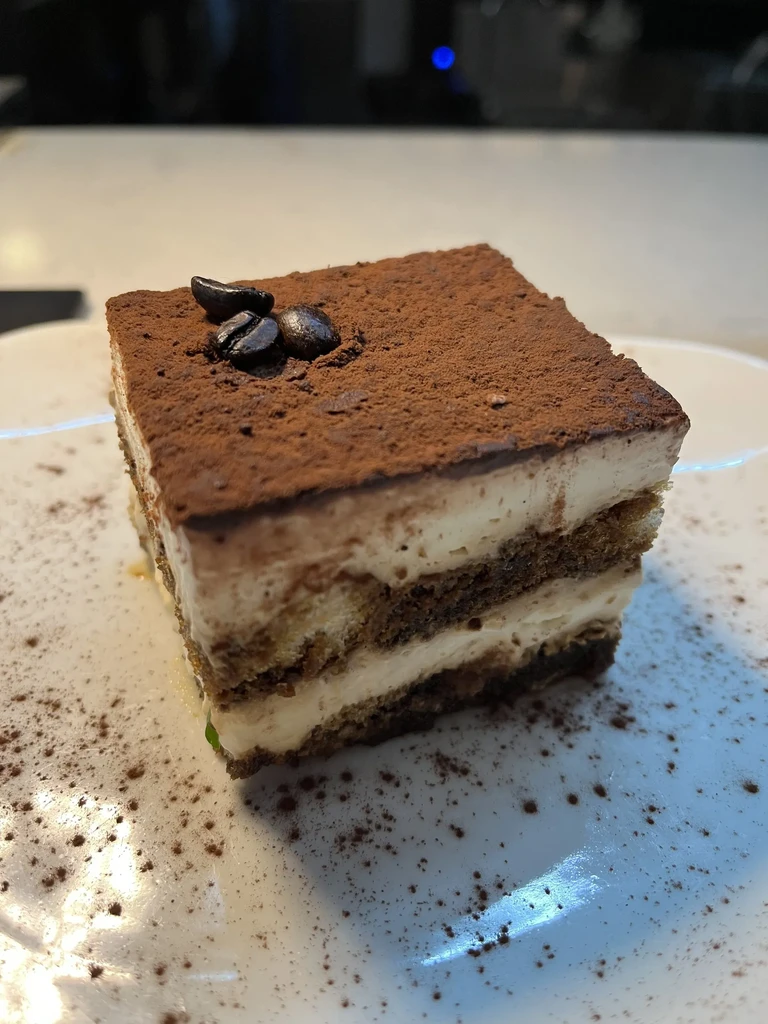 Layered Tiramisu slice with cocoa powder and coffee beans on a white plate.