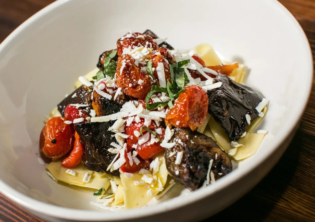 White bowl of pasta with roasted eggplant, tomatoes, cheese, and herbs.