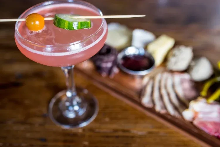 Pink cocktail garnished with tomato and cucumber on a charcuterie board.