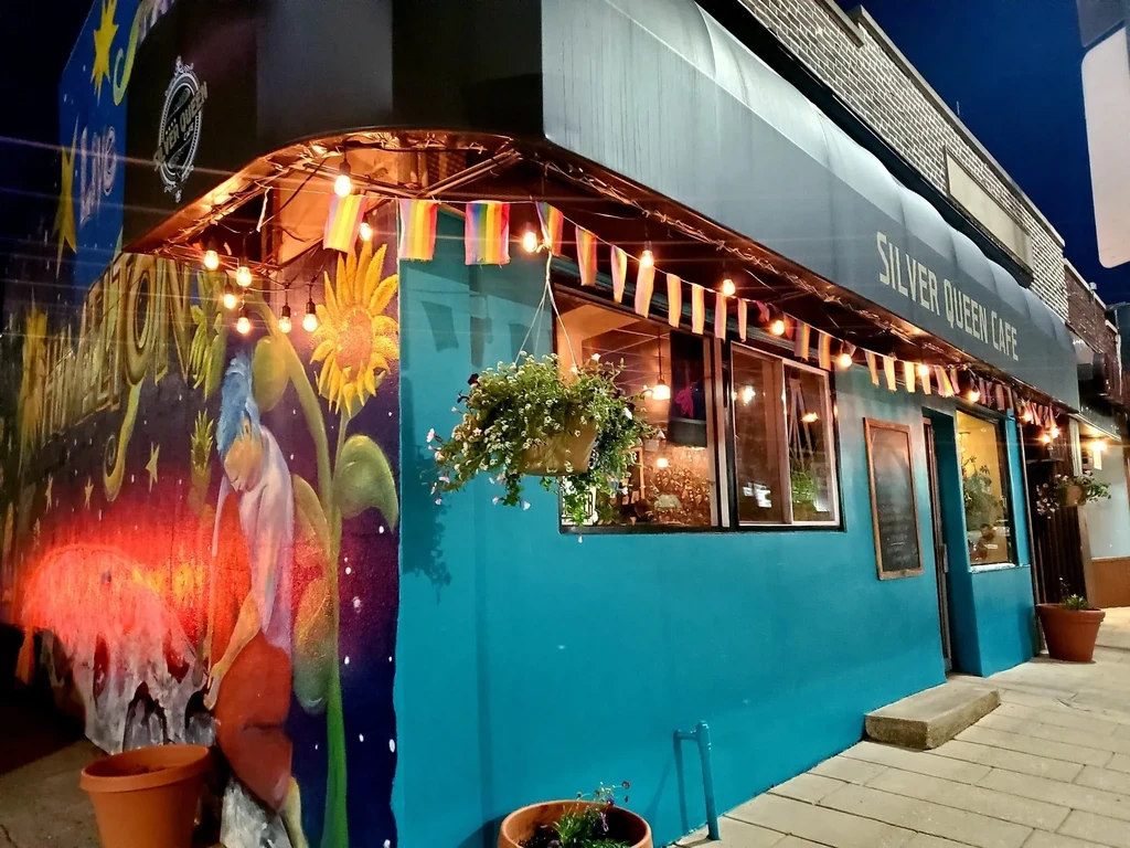 Silver Queen Cafe exterior at night: turquoise facade, colorful mural, rainbow flags.