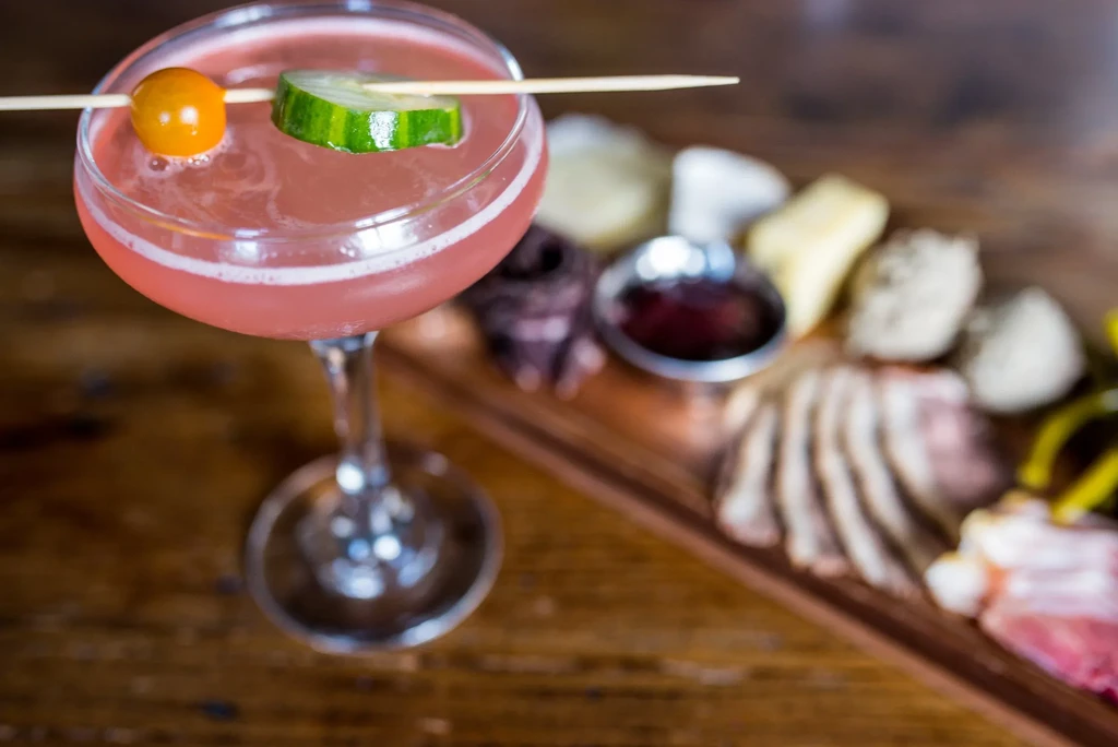 Pink cocktail with cherry and cucumber garnish, blurred charcuterie background.