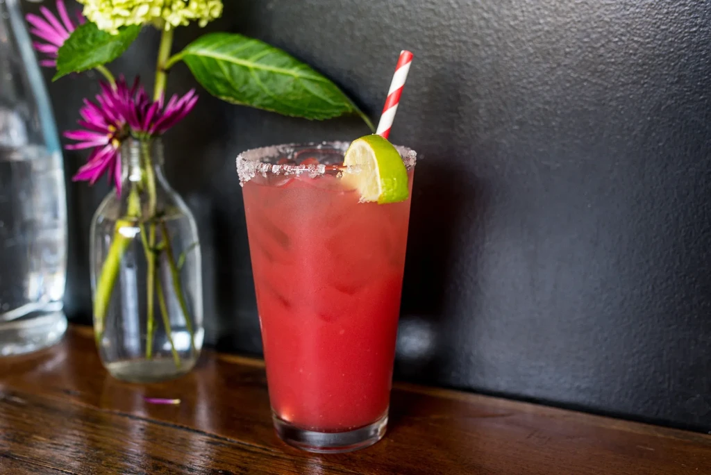 Red drink with lime, straw, and salted rim on a wooden table.