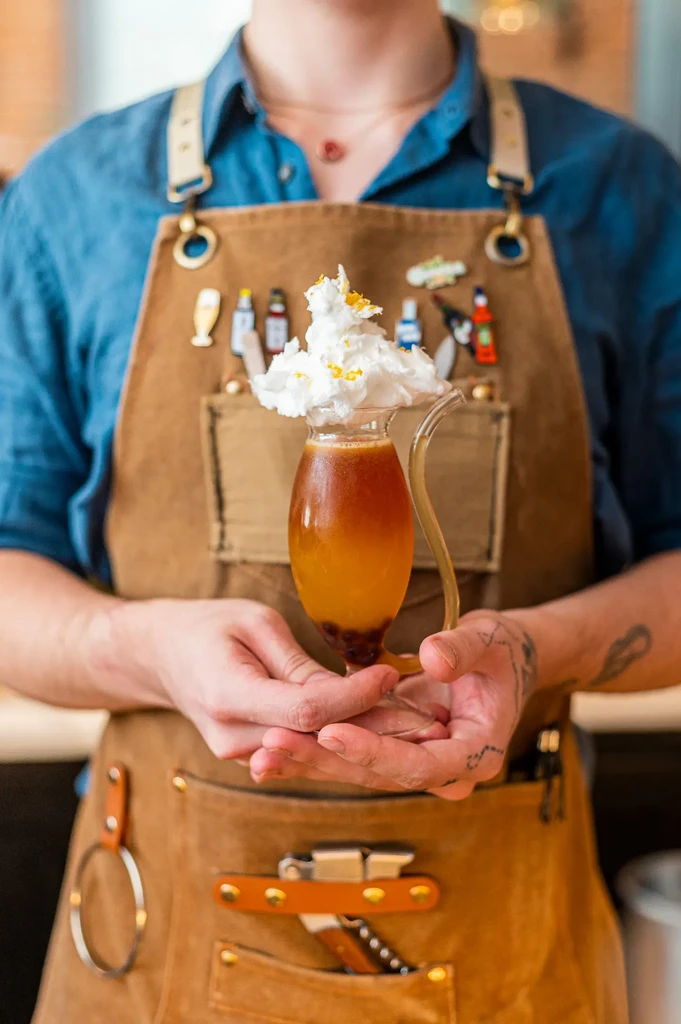 Bartender in apron with pins holds layered whipped cream cocktail.