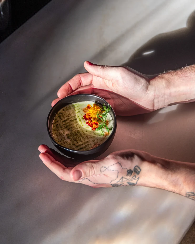 Creamy plated dish with green oil, edible flowers, held in dark bowl.