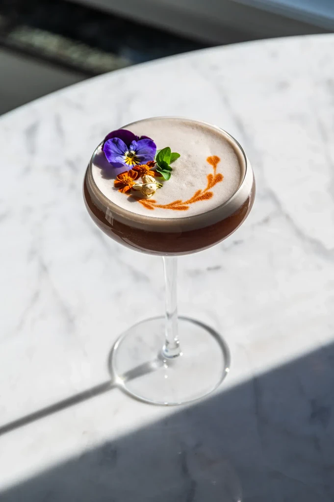 Dark cocktail in coupe glass with foamy top, flowers, and orange design.