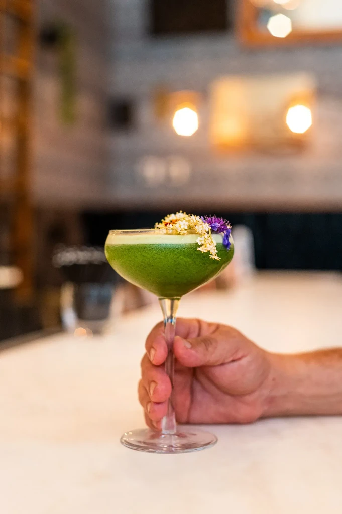 Hand holding green foamy cocktail in coupe glass, garnished with flowers.