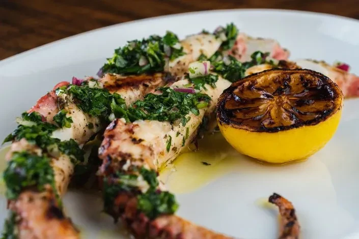 Grilled octopus with green chimichurri, charred lemon, on a white plate.