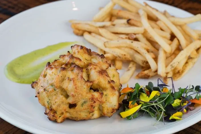 A fried patty, french fries, green sauce, and colorful garnishes on a plate.