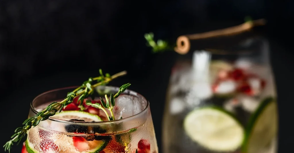Two cold drinks garnished with lime, pomegranate, thyme, dark background.