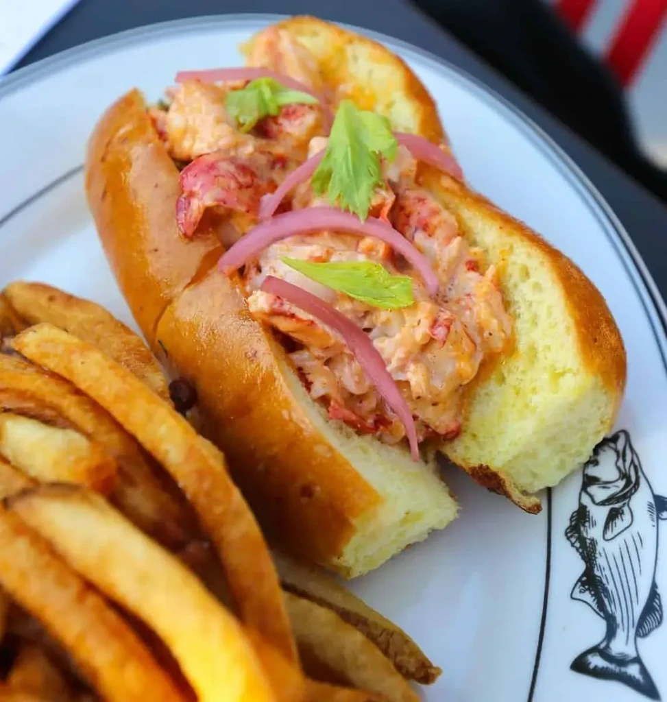 Lobster roll with pickled onions, herbs, and fries on fish-drawn plate.