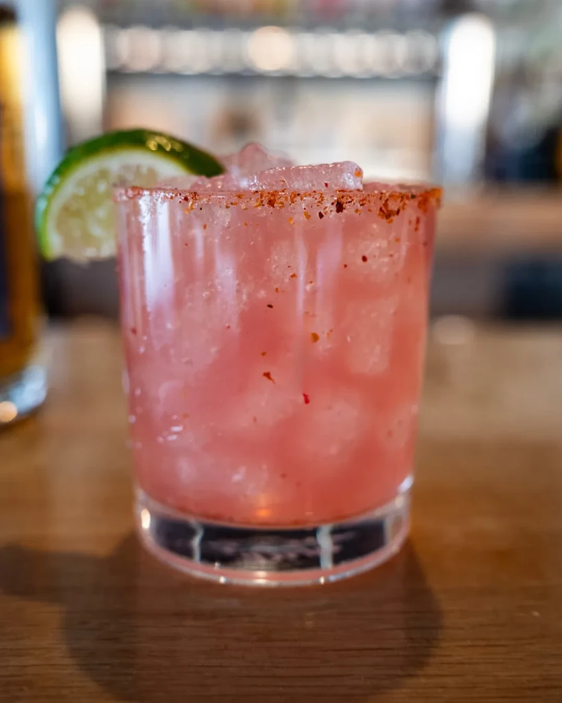 Frosty pink cocktail with lime and spiced rim on wooden surface.