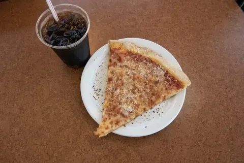 Pizza slice, soda with straw, on a speckled brown table.
