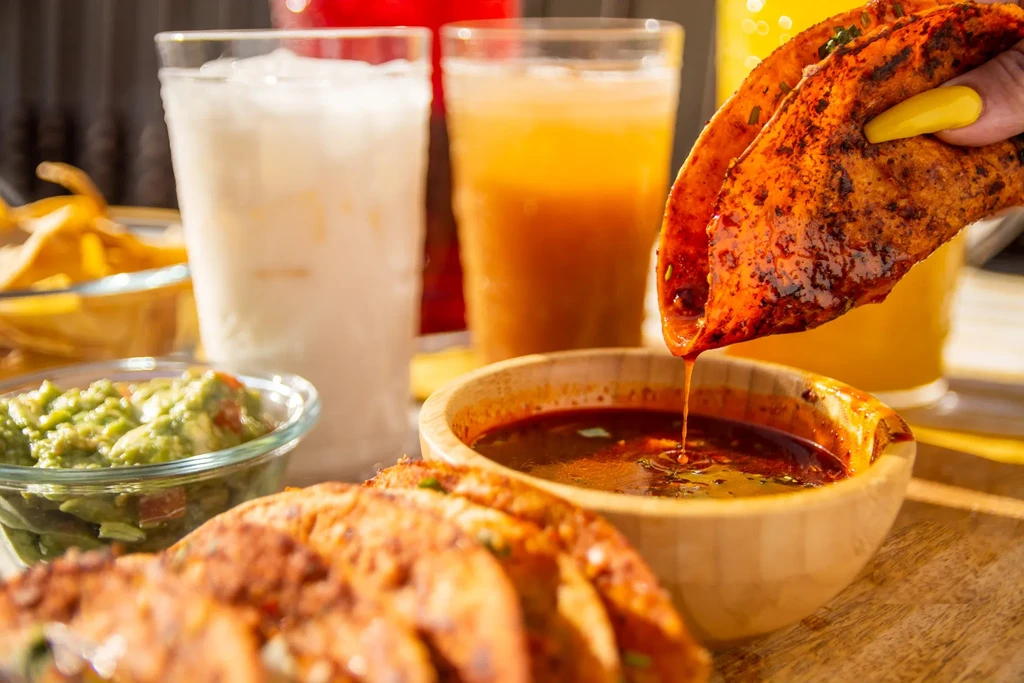 Vibrant birria tacos, one dipping into consome, with guacamole and drinks.