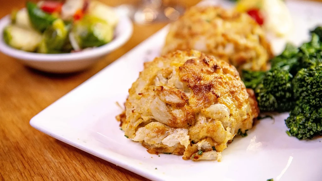Two crab cakes with broccoli and vegetables on a white plate.