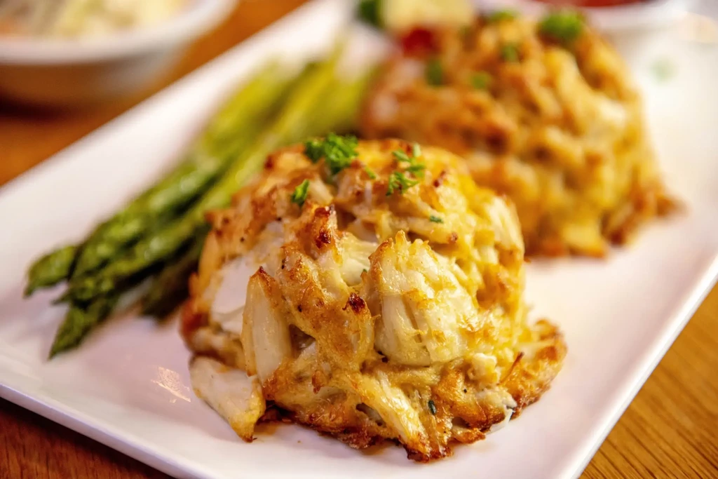 Two golden-brown crab cakes, topped with herbs, served with asparagus.
