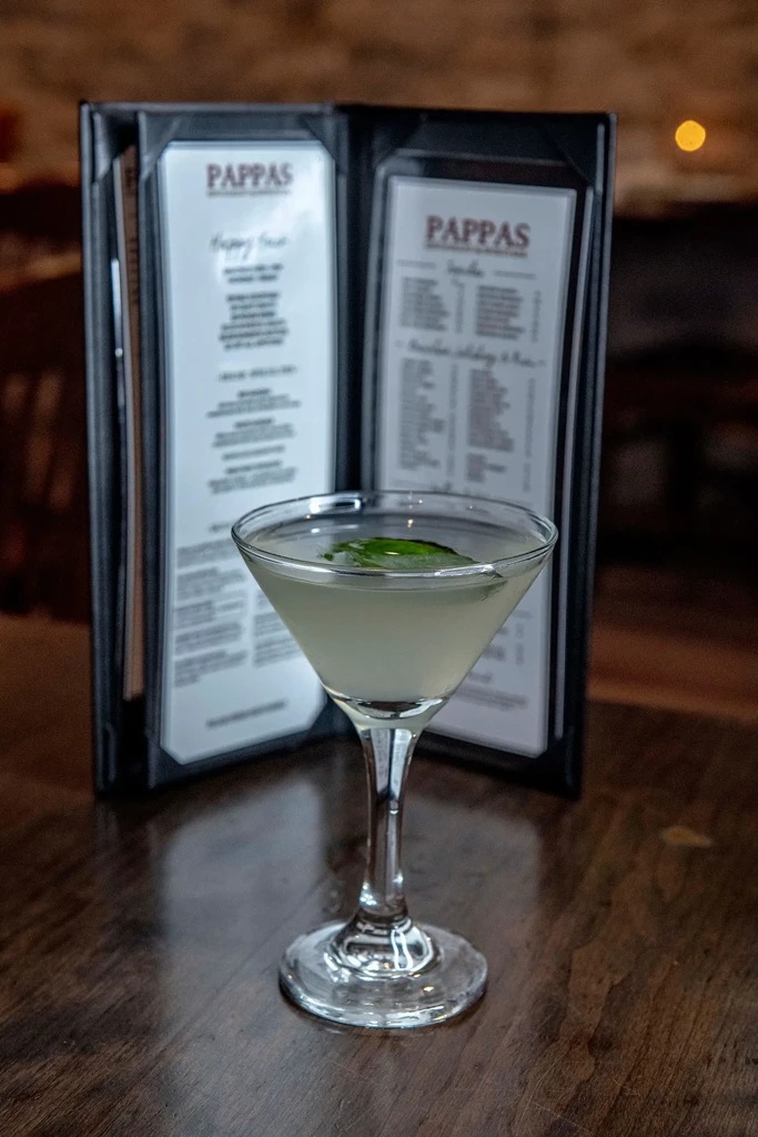 Martini glass with light cocktail and green leaves, blurred PAPPAS menu background.