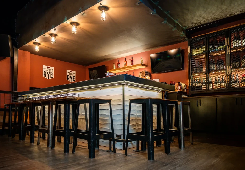 Industrial-style bar interior with long counter, stools, and liquor displays.