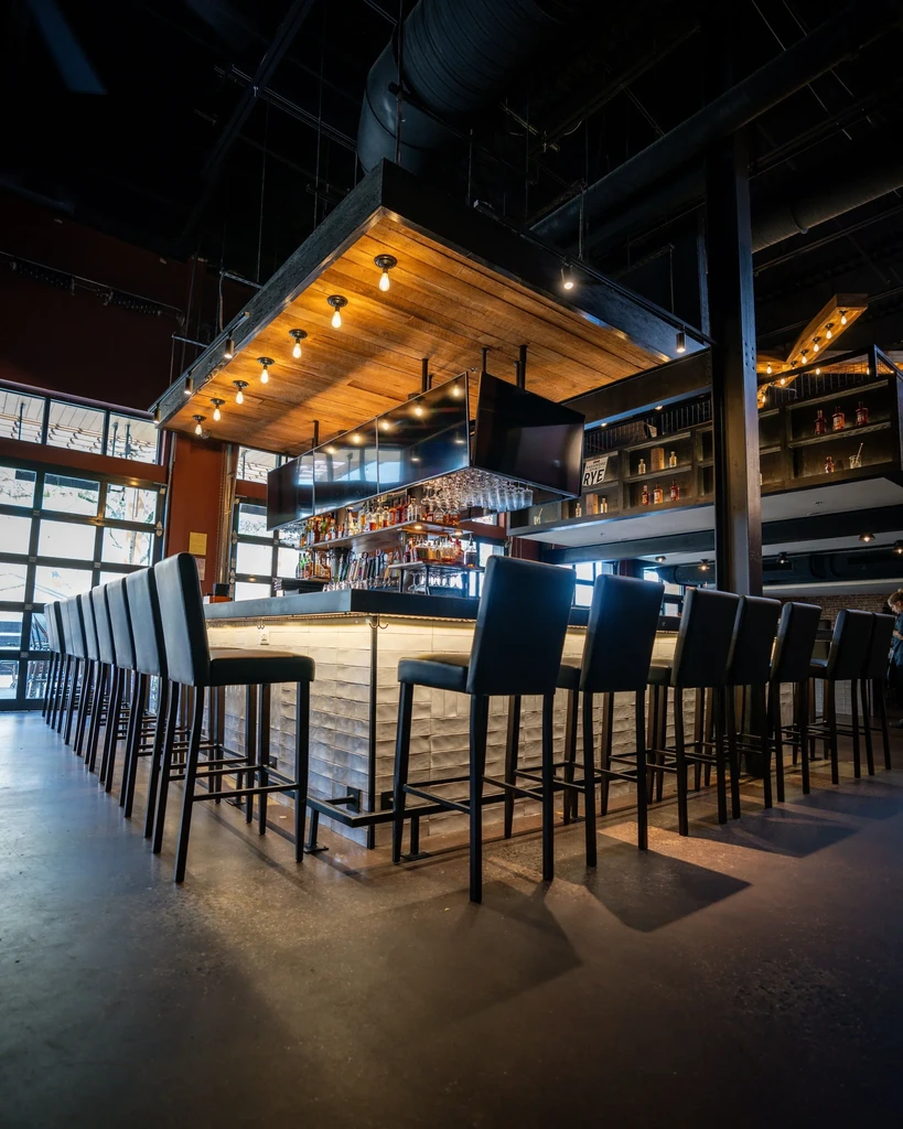 Modern bar with dark decor, high stools, glowing counter, and wooden ceiling.