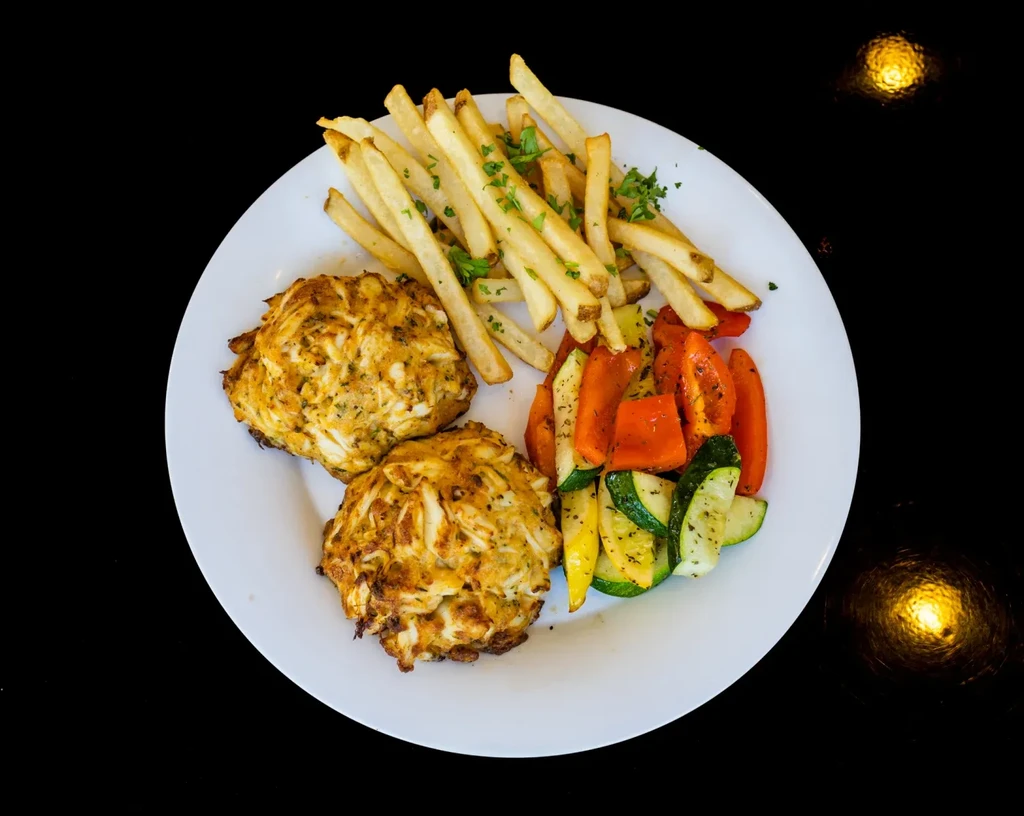 Two golden crab cakes, fries, and roasted vegetables on a white plate.