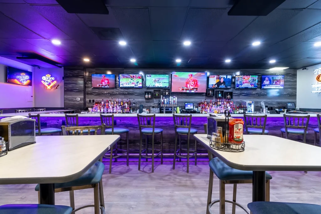 Modern bar interior with purple lighting, TVs showing sports, and neon signs.