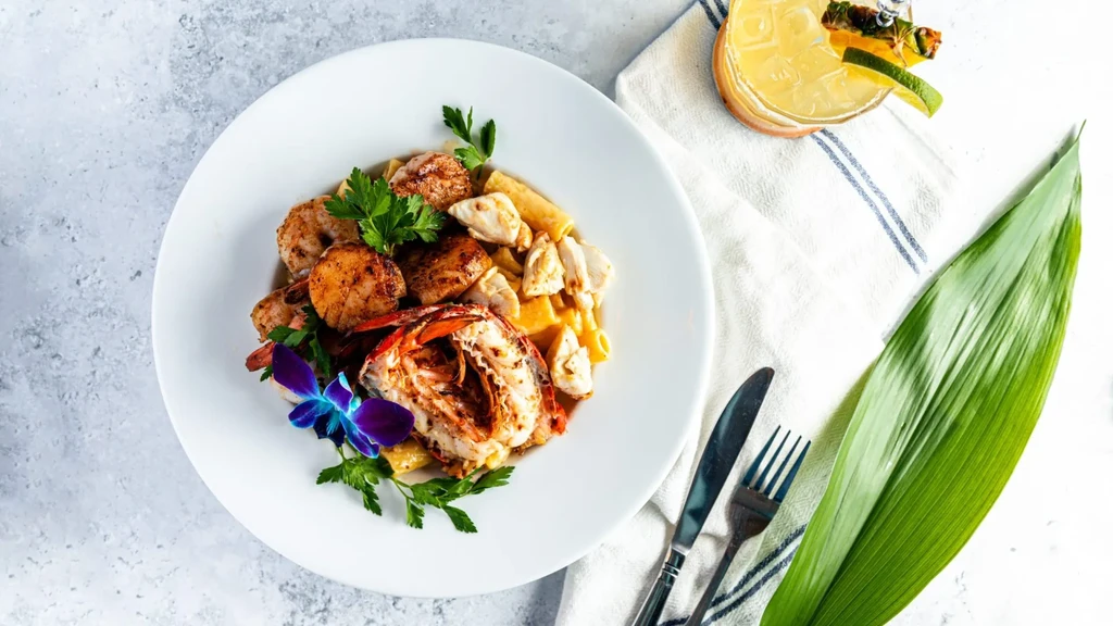 Gourmet lobster and scallop pasta with orchid garnish, cocktail, and tropical leaf.