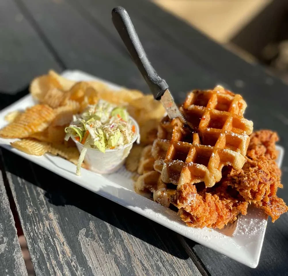 Chicken and waffles, coleslaw, and chips on a rustic wooden table.