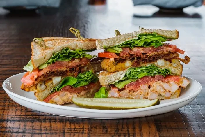 Stacked club sandwich with chicken, shrimp, bacon, lettuce, tomato, and pickle.