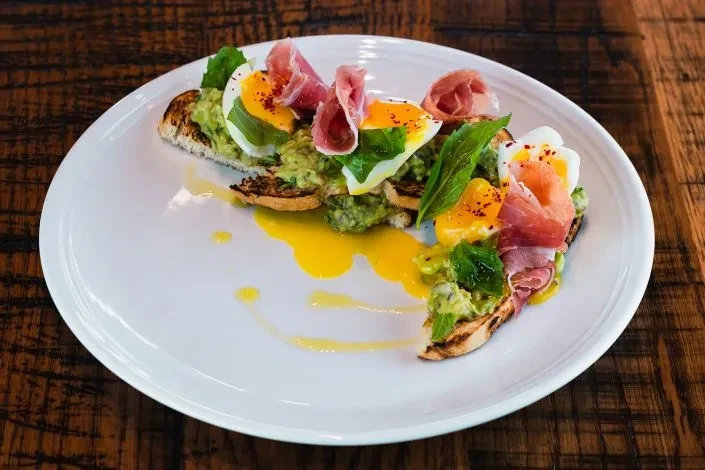 Artfully arranged avocado toast with runny eggs, prosciutto, greens, and spice.