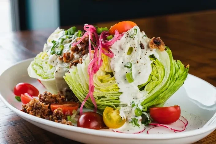 Deconstructed wedge salad with iceberg, creamy dressing, tomatoes, bacon, pickled onions.