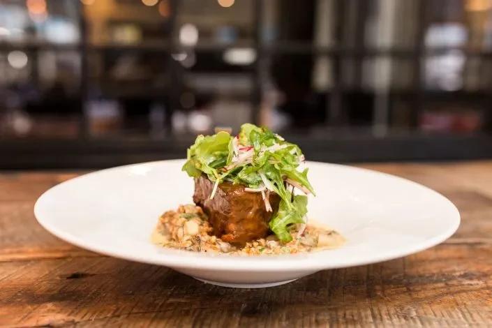 Braised meat, green salad, grains, and creamy sauce on white plate.