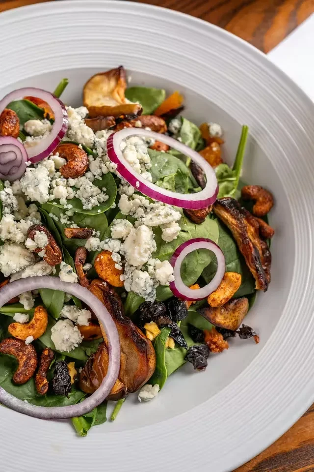 Spinach salad in white bowl with blue cheese, nuts, and dried fruit.