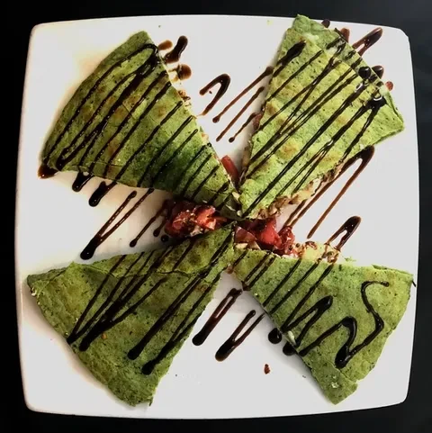 Four green crepes, strawberry filling, chocolate drizzle on white plate.